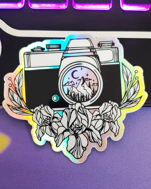 A silver and black holographic classic film camera waterproof vinyl sticker with a night time mountain scene reflecting in the lens.  Flowers and leaves surrounding the camera.  Perfect for camera lovers and photographers of nature.