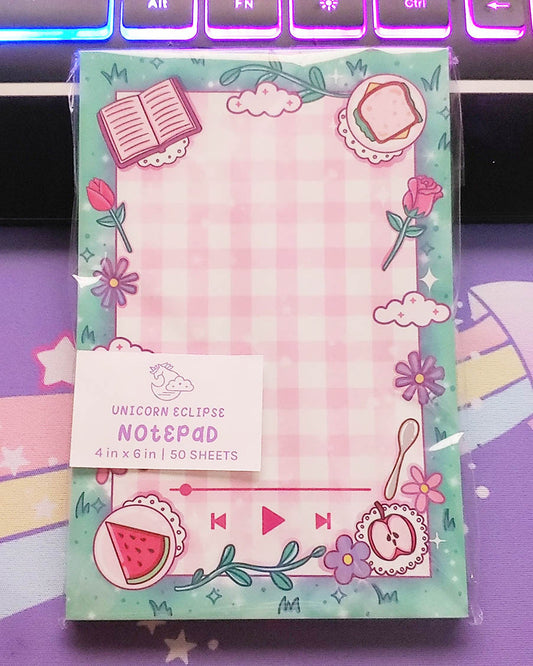 A 50 sheet non-sticky notepad measuring 4" by 6" featuring a picnic blanket sitting on green grass.  On the picnic blanket lies a book, sandwich on a plate, a rose, two clouds, 4 misc flowers, a spoon, an apple on a doily, a watermelon, and two vines.  There are also mp3 player icons at the bottom.