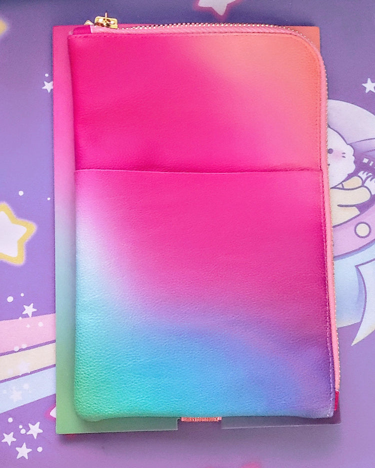 A green, blue, purple, pink, and coral ombre vegan leather folio planny pack pouch with gold toned zipper is laying flat against a purple astronaut kitty in a spaceship large mousepad.