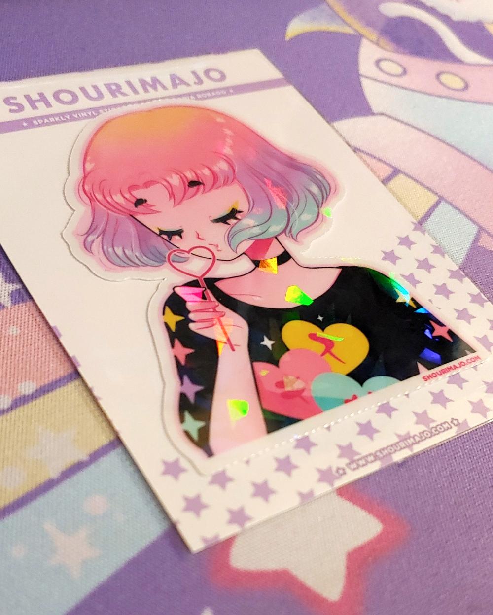 A young lady/adult/woman with yellow, pink, purple, and blue pastel ombre hair is blowing bubbles through a heart wand.  Her eyes are closed and she has blue and yellow eye shadow.  She is wearing a black choker and a black long sleeve shirt with yellow, pink, purple, and blue stars on the sleeves.  The shirt also has yellow, blue, and pink hearts on the front.  This digital art piece is print on a broken holographic vinyl sticker.  The sticker is attached to a white purple starred backing card.
