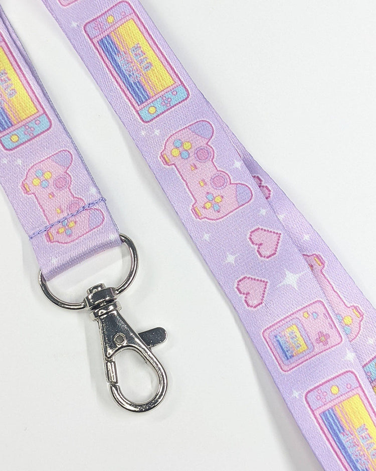A pastel purple lanyard featuring pink playstation gaming controllers. Each controller has pastel yellow & blue buttons, pink joysticks, and purple grippers.  There are also pastel pink, yellow, blue, & purple nintendo switches that read "game over".  Another item is a pastel pink, yellow, and blue game boy that also reads, "game over".  There are white sparkles and pink pixel hearts.  This lanyard has a metal clasp.