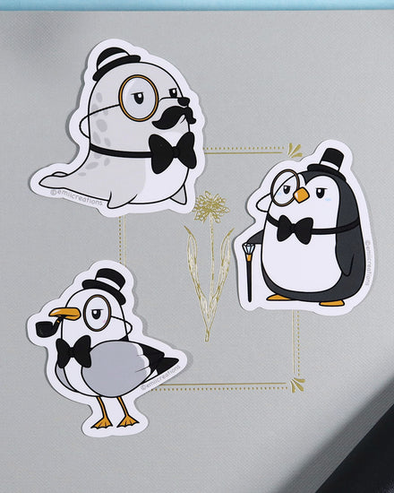 A grey gray chibi seal with a gold toned monocle, black and white bully hat, black mustache, and black bow tie vinyl sticker. Design by small business Emii Creations.  This image also features other styles including a pigeon and a penguin wearing a top hat.