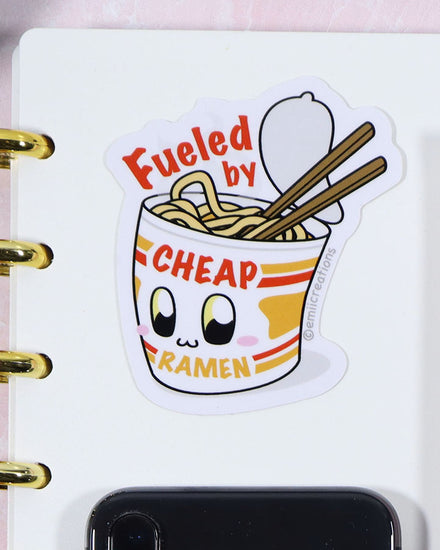 A kawaii chibi vinyl sticker featuring a cup of instant lunch ramen noodles. The image shows that the pack on instant lunch noodles is open and you can see the noodles. There are chopsticks sticking into the noodles. The packaging has a face on it and it's blushing and smiling. The sticker reads "Fueled by Cheap Noodles" The sticker is designed by small business Emii Creations