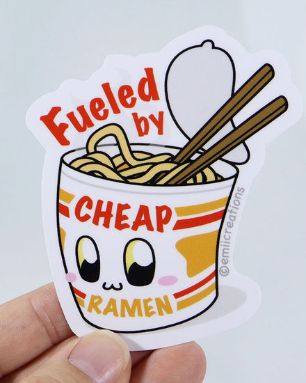 A kawaii chibi vinyl sticker featuring a cup of instant lunch ramen noodles.  The image shows that the pack on instant lunch noodles is open and you can see the noodles.  There are chopsticks sticking into the noodles.  The packaging has a face on it and it's blushing and smiling.  The sticker reads "Fueled by Cheap Noodles"  The sticker is designed by small business Emii Creations