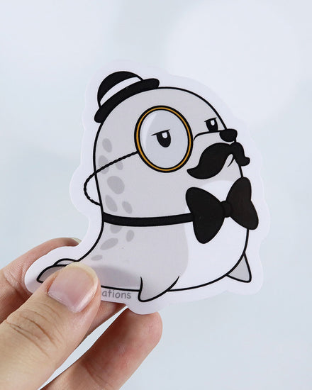 A grey gray chibi seal with a gold toned monocle, black and white bully hat, black mustache, and black bow tie vinyl sticker.  Design by small business Emii Creations