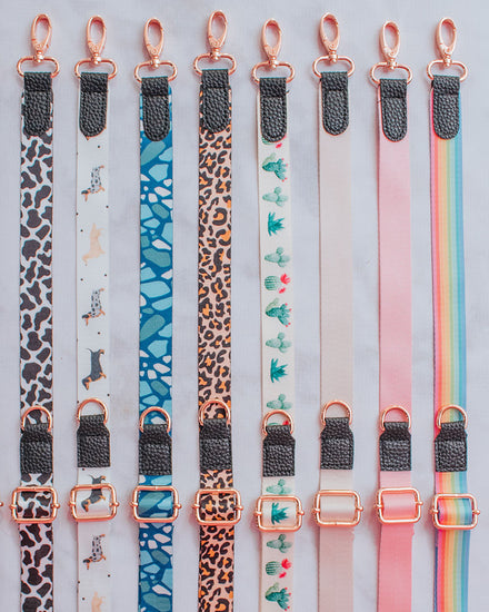 Eight build-your-own crossbody straps laying flat next to each other in the following order: black and white cow print, black, white, and yellow dachshund print on white background, blue and white desert floor print, leopard print, green, pink, and white cactus print, beige, pastel pink, and sassy stripes rainbow print.