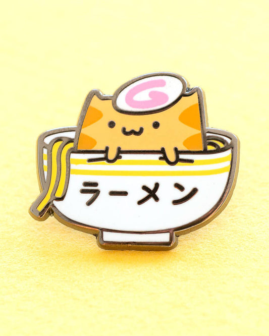 A kawaii orange cat sitting in a bowl of ramen noodles with an egg on it's head on an enamel pin.  Designed by robot dance battle