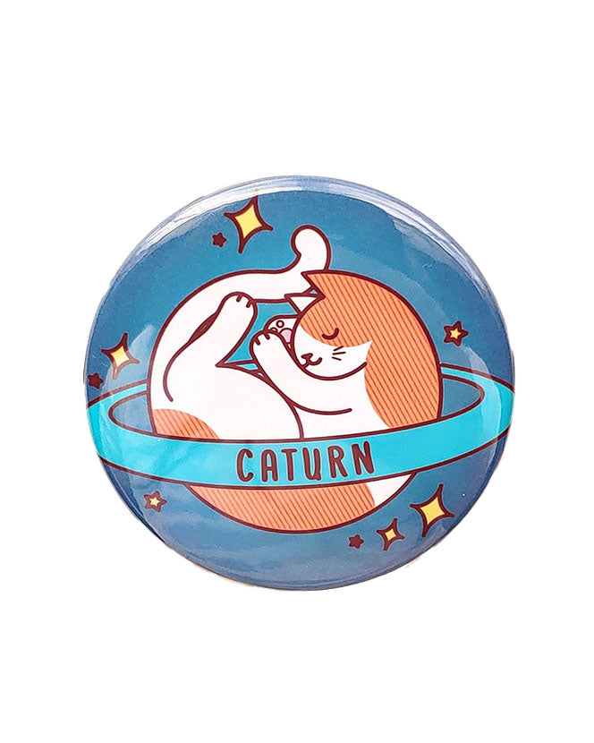An orange and white saturn style planet with a ring that says caturn on the ring on a blue background with yellow stars. The cat is sleeping. It is a magnet.