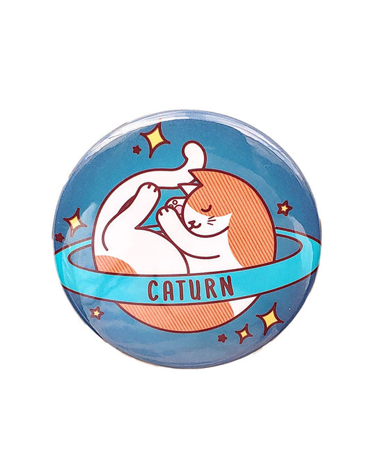 An orange and white saturn style planet with a ring that says caturn on the ring on a blue background with yellow stars. The cat is sleeping. It is a magnet.