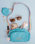 A build-your-own sassy woof example featuring blue 2 compartment crossbody bag pouch with a Sassy Stripes pastel rainbow strap and gold-toned clasps, attached near the top is a blue 3.45" (inches) circular treat pouch. The photo shows how the crossbody bag can be used like a purse as well as a waste bag holder.