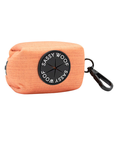 A SASSY WOOF Apple Cider orange rectangular 3" pouch with black zipper and black clasp. The rubber part where the waste bags come out in black and reads "SASSY WOOF" in white writing.