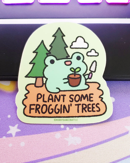 A cute kawaii smiling frog froggie holding a gardening spade and a plant with dirt and a leaf sitting in camp dirt with two pine trees behind him and two clouds in the sky on a vinyl sticker. The sticker reads "Plant some froggin' trees." Designed by small business robot dance battle